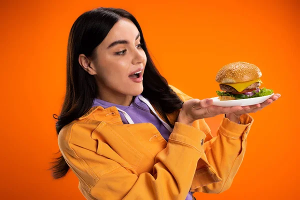 Amazed woman in stylish jacket looking at tasty burger with cheese and meat isolated on orange - foto de stock