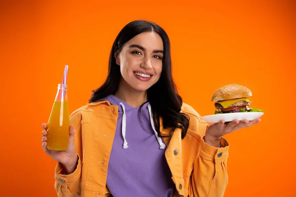Счастливая женщина с вкусным бургером и бутылкой свежего лимонада глядя на камеру изолированы на оранжевый — стоковое фото