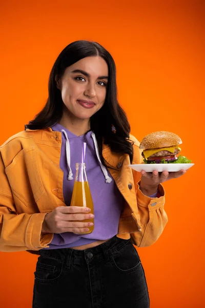 Стильная женщина с вкусным бургером и свежим лимонадом улыбаясь в камеру изолированы на оранжевый — стоковое фото