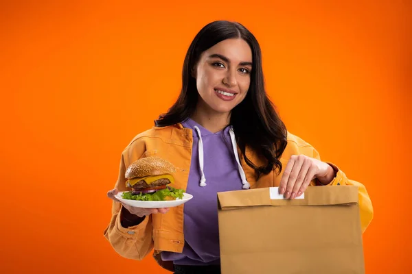 Happy brunette woman holding paper bag and delicious burger isolated on orange - foto de stock