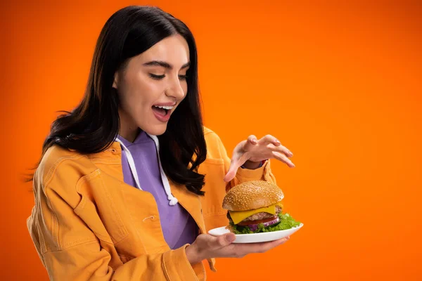 Hungry woman holding delicious burger with cheese and meat isolated on orange — Stock Photo