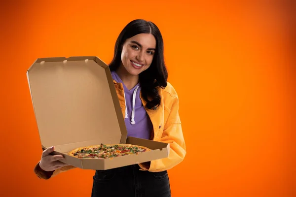 Joyful woman looking at camera while holding carton box with tasty pizza isolated on orange — Stockfoto
