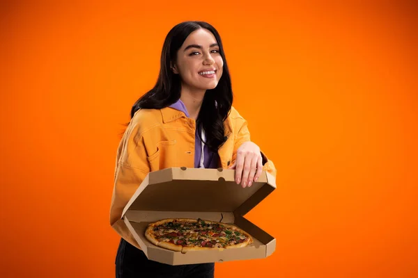 Smiling brunette woman showing tasty pizza in carton box while looking at camera isolated on orange — Photo de stock