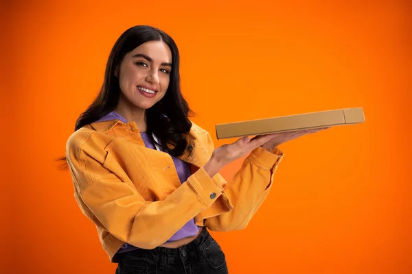 Cheerful woman in trendy jacket holding pizza box and looking at camera isolated on orange — Photo de stock