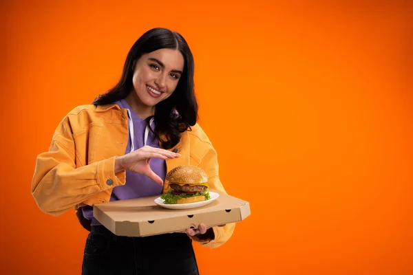 Brunette woman with delicious burger and pizza box smiling at camera isolated on orange - foto de stock