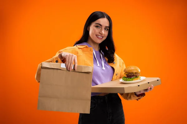 Усміхнена жінка тримає смачний бургер і паперовий мішок, дивлячись на камеру ізольовано на помаранчевому — стокове фото