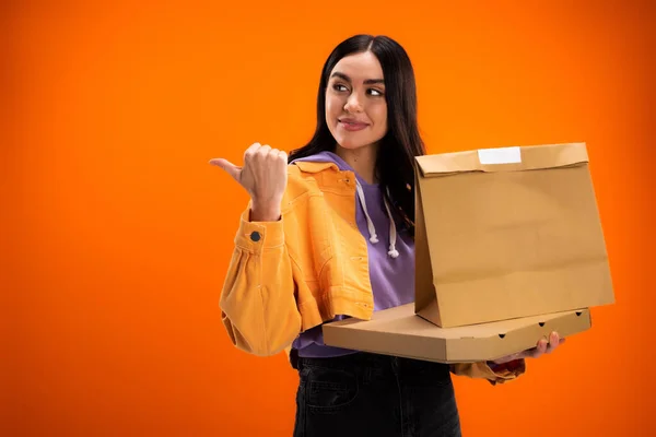 Young woman with paper bag and pizza box looking away and pointing with thumb isolated on orange — Foto stock