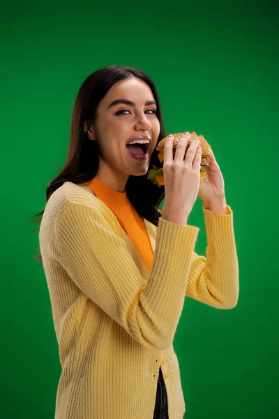 Joyful woman looking at camera while opening mouth near tasty burger isolated on green - foto de stock