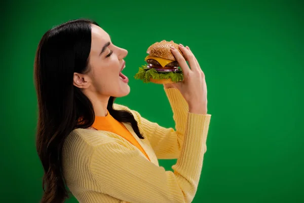 Side view of young and hungry woman opening mouth near burger with cheese and meat isolated on green — Stock Photo