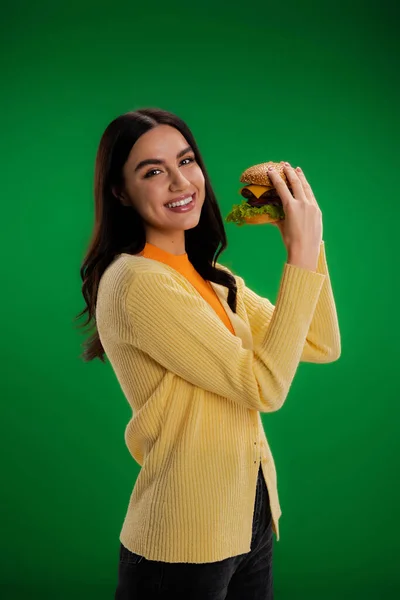 Веселая брюнетка смотрит в камеру, держа в руках вкусный бургер, изолированный на зеленом — стоковое фото
