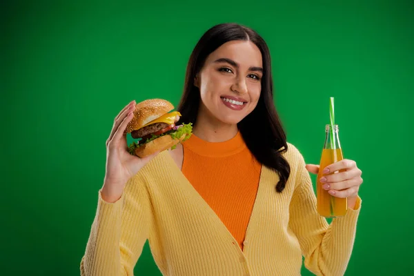 Happy brunette woman holding delicious burger and bottle of fresh juice isolated on green - foto de stock