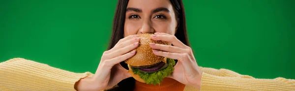 Young and hungry woman eating hamburger and looking at camera isolated on green, banner — Foto stock