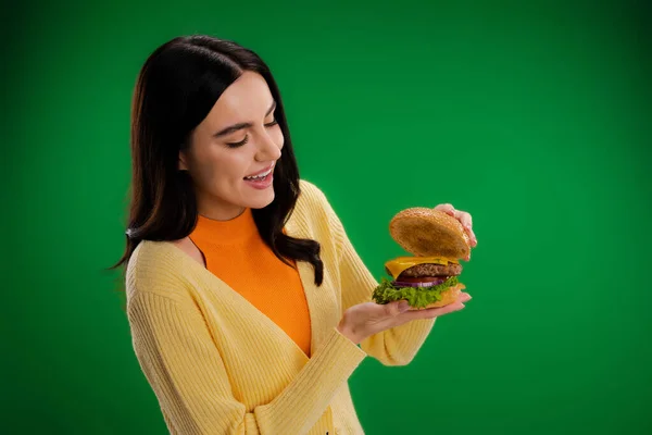 Здивована жінка дивиться на смачний бургер з сиром і м'ясом ізольовано на зеленому — стокове фото