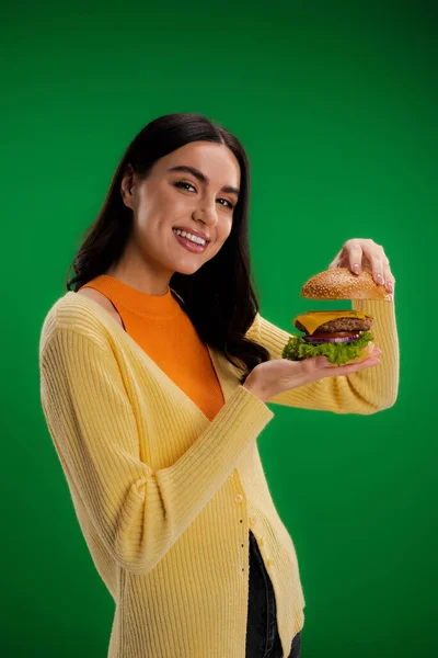 Young brunette woman showing delicious burger while smiling at camera isolated on green — Photo de stock