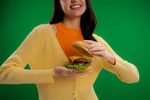 Обрезанный вид улыбающейся женщины, держащей бургер с мясом и сыром с овощами изолированы на зеленый — стоковое фото