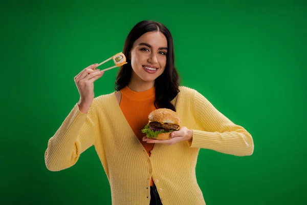 Happy young woman holding burger and chopsticks with sushi roll isolated on green — Fotografia de Stock