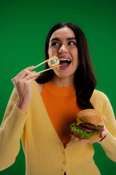 Pleased woman holding burger and eating fresh sushi roll with chopsticks isolated on green - foto de stock