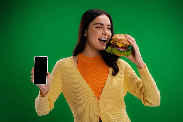 Young woman with delicious burger showing smartphone with blank screen isolated on green — Stock Photo