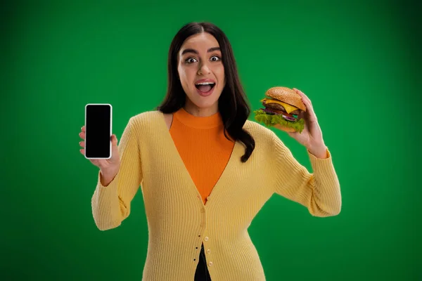 Изумленная женщина держит вкусный бургер и мобильный телефон с чистым экраном изолированы на зеленый — стоковое фото