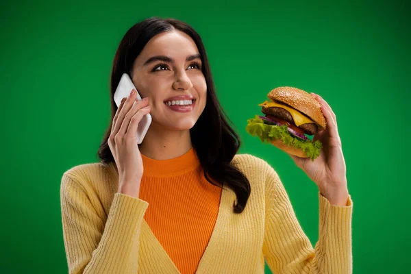 Щаслива молода жінка тримає смачний бургер і розмовляє на смартфоні ізольовано на зеленому — стокове фото