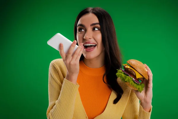 Щаслива жінка тримає смачний бургер і відправляє голосове повідомлення на смартфон ізольовано на зеленому — стокове фото