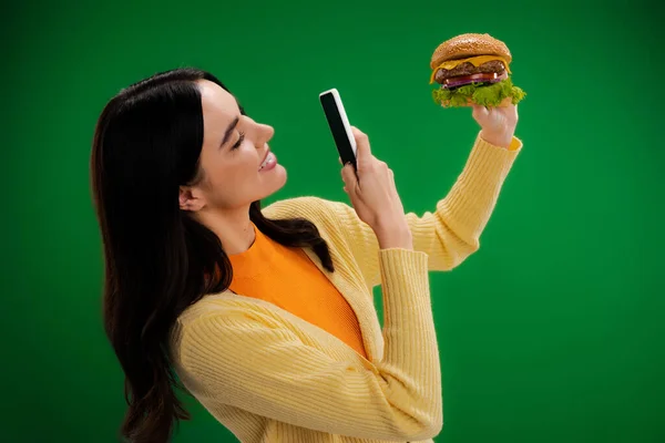 Smiling woman taking photo of delicious burger on smartphone isolated on green — Photo de stock