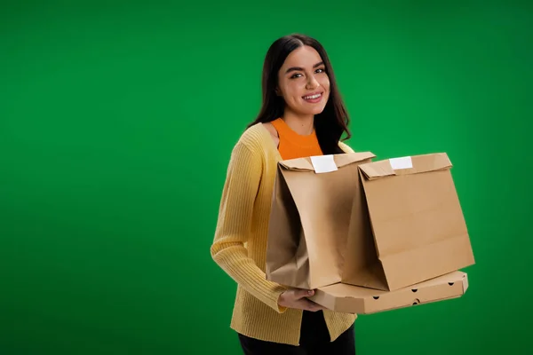 Happy brunette woman holding paper bags and pizza box while looking at camera isolated on green - foto de stock