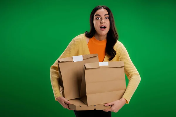 Verblüffte Frau mit Lebensmittelpaketen blickt in Kamera isoliert auf Grün — Stockfoto