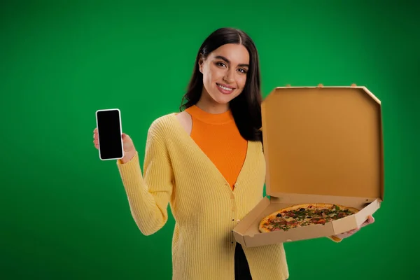 Smiling woman with box of delicious pizza showing smartphone with blank screen isolated on green — Photo de stock