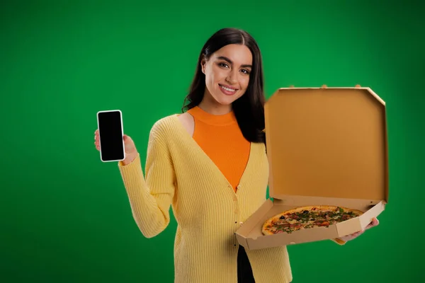 Smiling woman with box of delicious pizza showing smartphone with blank screen isolated on green — Stock Photo