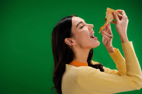 Вид сбоку молодой и голодной женщины, которая ест кусок пиццы, изолированный на зеленом — стоковое фото