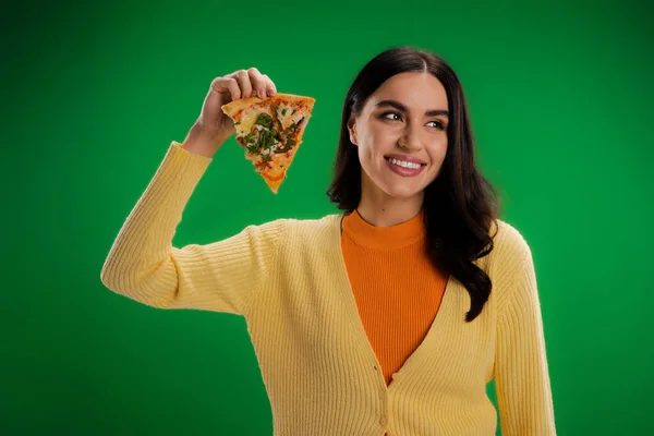 Cheerful woman in orange turtleneck and yellow jumper holding piece of pizza and looking away isolated on green - foto de stock