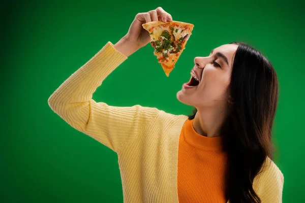 Mujer joven en jersey amarillo sosteniendo deliciosa pizza cerca de la boca abierta aislado en verde - foto de stock