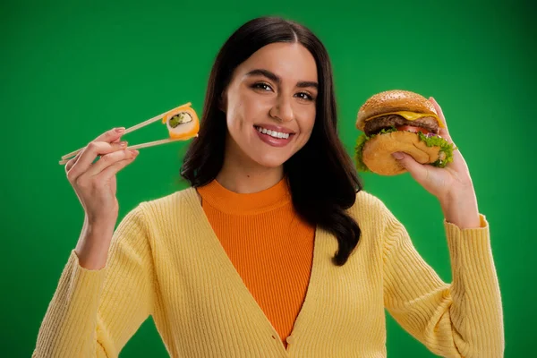 Cheerful woman holding tasty burger and fresh sushi while looking at camera isolated on green — Stock Photo