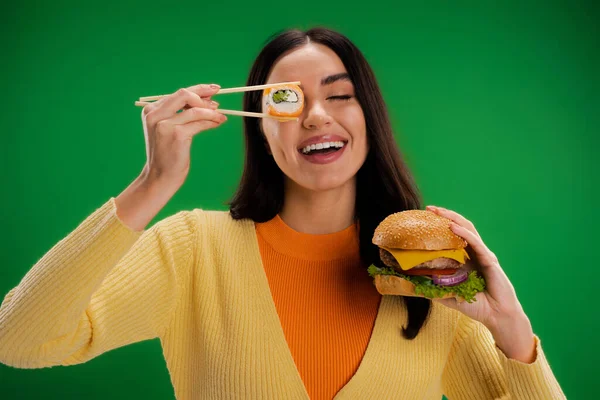 Cheerful woman with tasty burger holding sushi roll near eye isolated on green - foto de stock