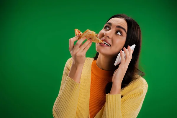 Хвилююча жінка їсть піцу під час розмови на смартфоні і дивиться вгору ізольовано на зеленому — стокове фото
