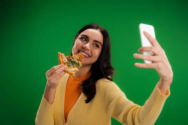 Cheerful woman biting piece of pizza and taking selfie on smartphone isolated on green — Foto stock