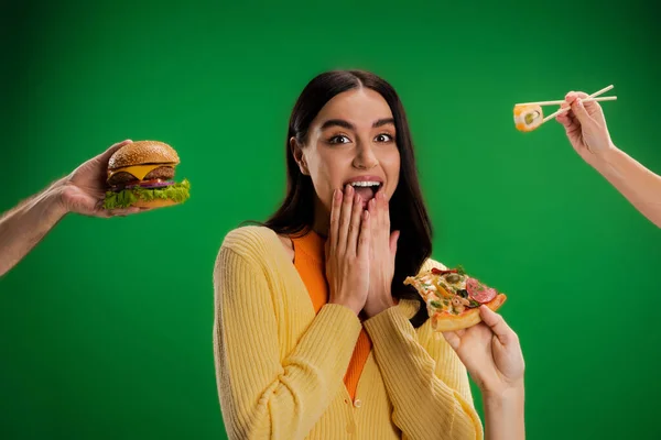 Здивована жінка, що покриває рот руками біля людей, пропонуючи різні продукти харчування ізольовані на зеленому — стокове фото