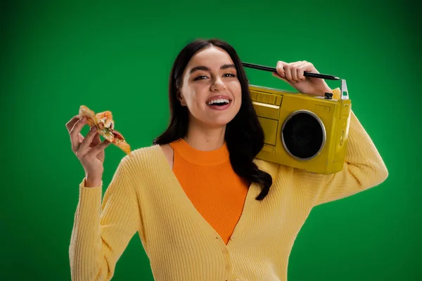 Joyful woman with piece of delicious pizza and boombox looking at camera isolated on green - foto de stock