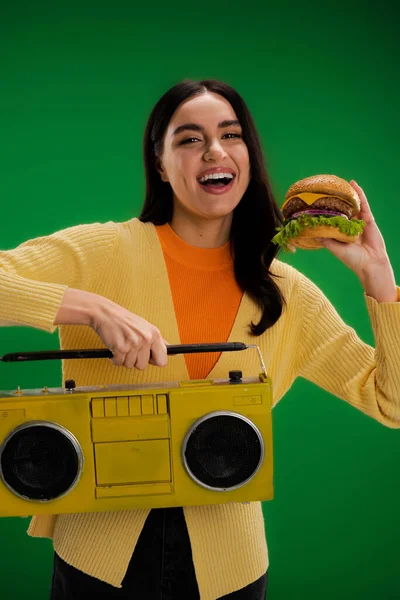 Смеющаяся женщина держит вкусный бургер и бумбокс, глядя на камеру, изолированную на зеленый — стоковое фото