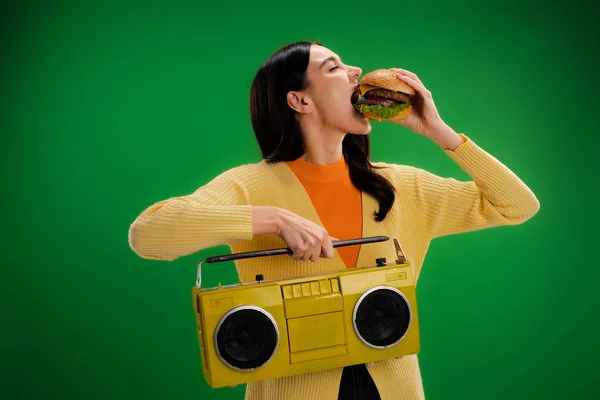 Young and hungry woman with boombox eating tasty burger isolated on green — Photo de stock
