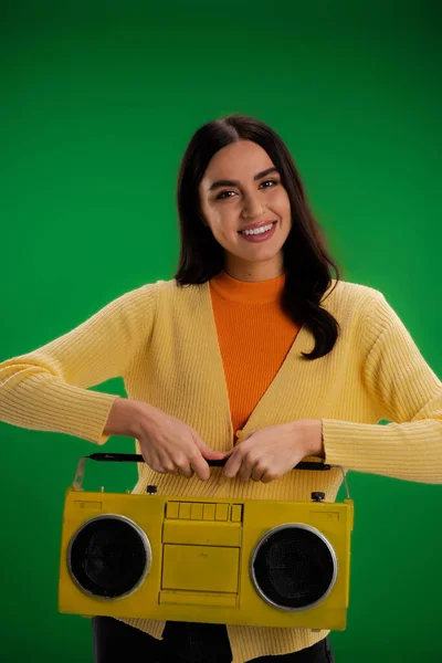 Brunette woman in yellow jumper and orange turtleneck holding boombox and looking at camera isolated on green - foto de stock