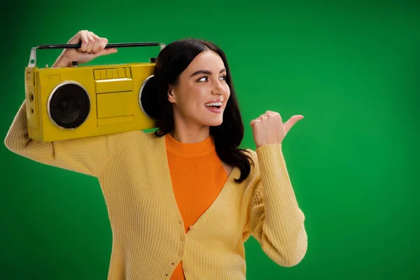Smiling woman with vintage boombox looking away and pointing with thumb isolated on green — Stock Photo