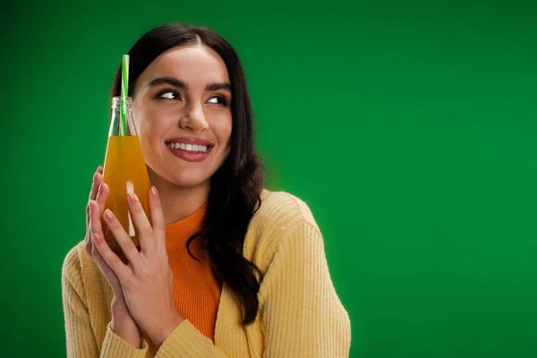 Радостная брюнетка женщина держит бутылку натурального лимонада и смотрит в сторону изолированы на зеленый — стоковое фото