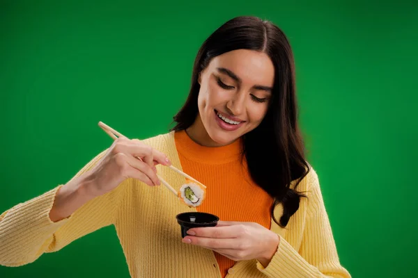 Smiling brunette woman holding sushi roll near sauce bowl isolated on green — Stock Photo