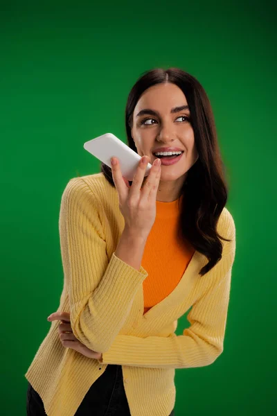 Cheerful brunette woman in orange turtleneck and yellow jumper recording voice message on smartphone isolated on green — Stock Photo