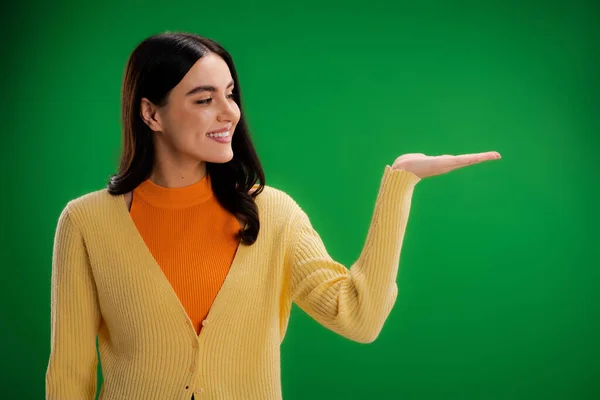 Smiling woman in orange turtleneck and yellow jumper pointing with hand isolated on green — Stockfoto