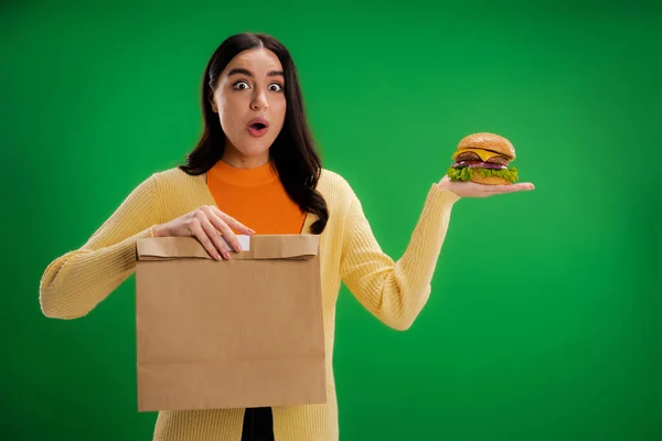 Amazed woman with tasty burger and paper bag looking at camera isolated on green - foto de stock