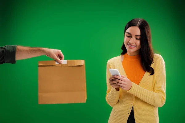 Smiling brunette woman using smartphone near delivery man with paper bag isolated on green — Stock Photo