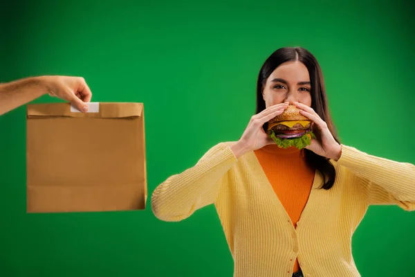 Голодна жінка їсть смачний бургер біля чоловіка з упаковкою їжі ізольовано на зеленому — стокове фото